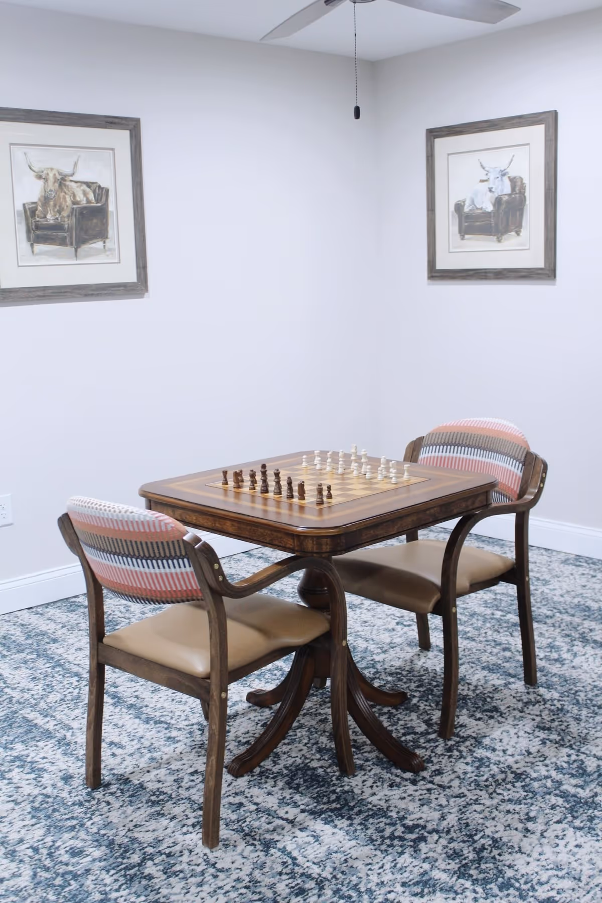 A small room with a wooden chess table set up for a game, surrounded by two wooden chairs with patterned cushions. The room has light-colored walls with two framed pictures of cows sitting on chairs, a ceiling fan above, and a blue patterned carpet.