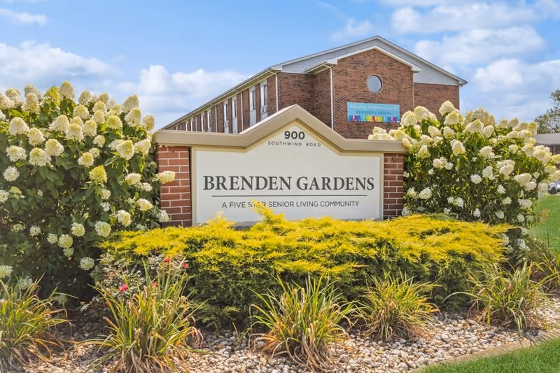 Entrance sign for Brenden Gardens in front of a brick senior living building surrounded by shrubs and flowers.