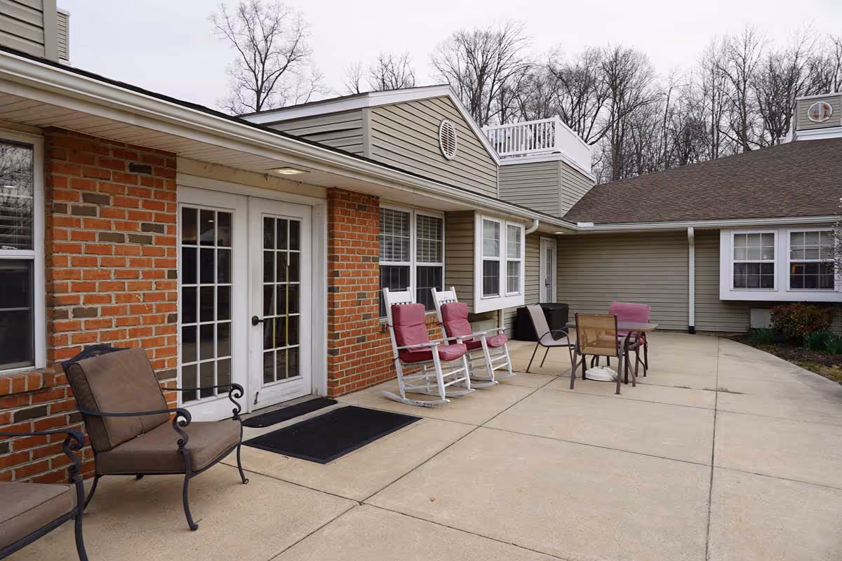 Outdoor patio area at Celebration Villa of Exeter featuring cushioned chairs, two white rocking chairs with pink cushions, a small table with chairs, and a brick and siding building exterior with windows and French doors.