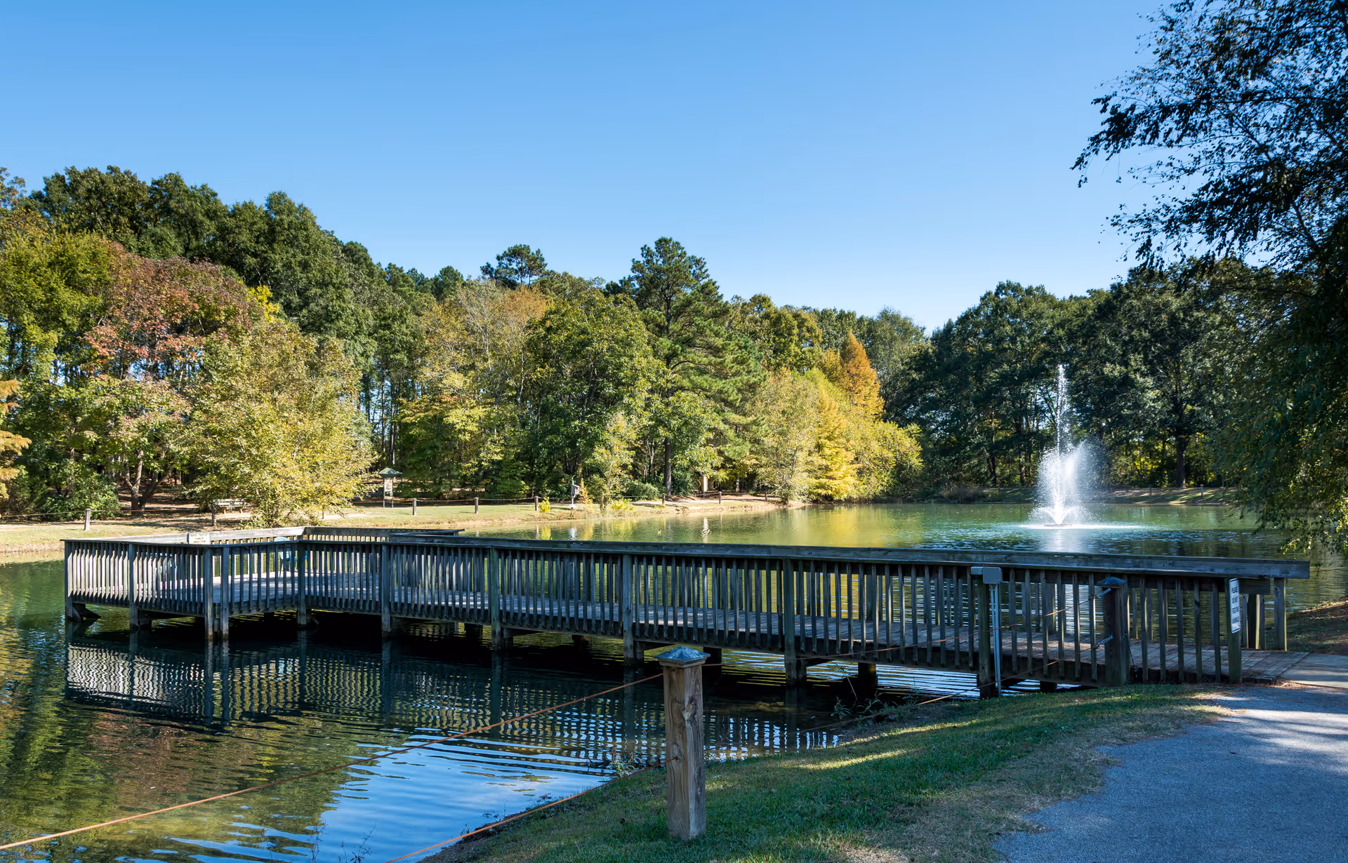 A wooden pier extending over a calm pond with a water fountain in the background, surrounded by green trees under a clear blue sky.