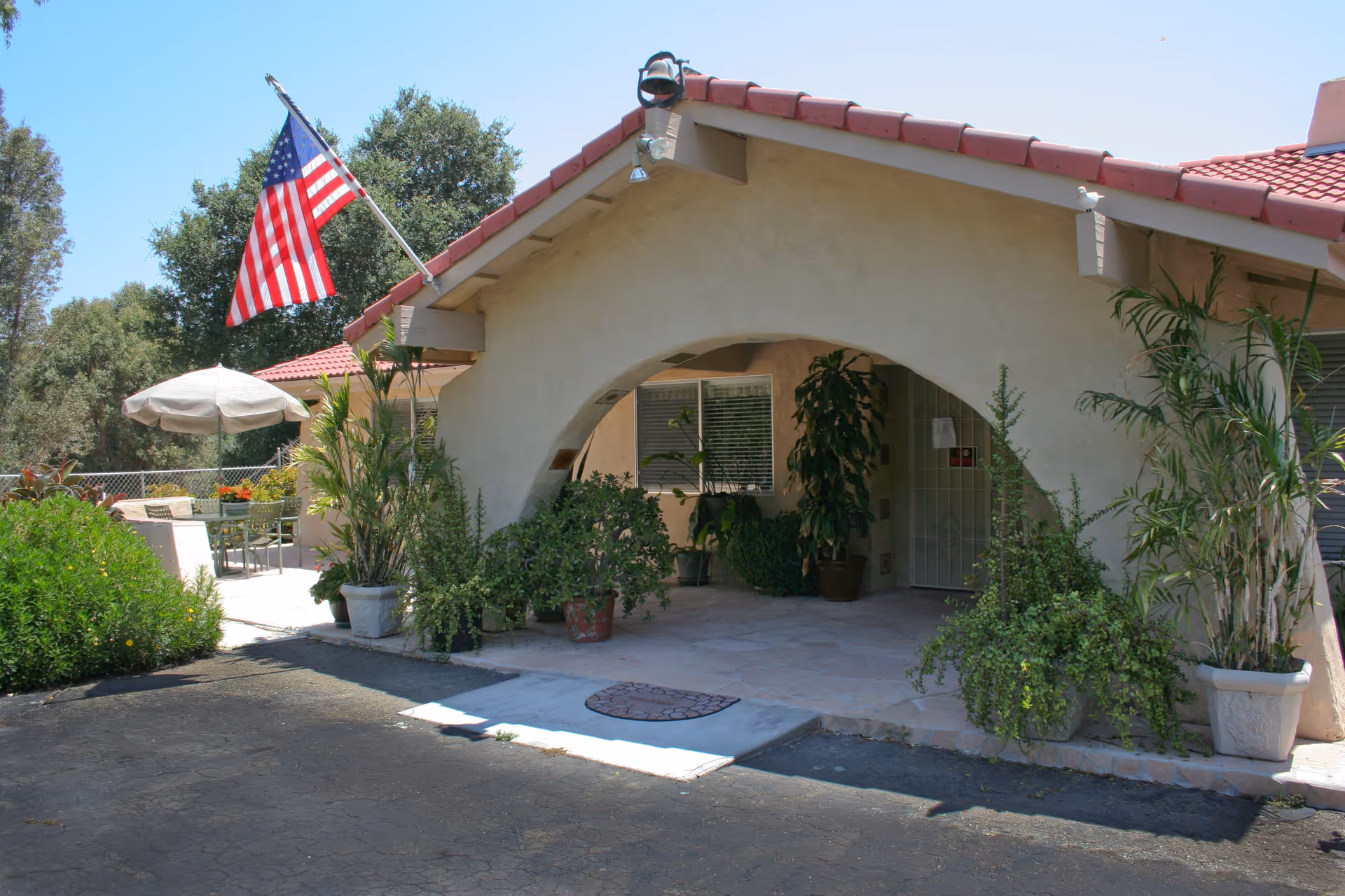Exterior view of a residential care facility with a beige stucco building featuring a red tile roof and an arched entrance. Several potted plants are placed around the entrance. An American flag is mounted on the building, and there is an outdoor seating area with a table and umbrella visible to the left.