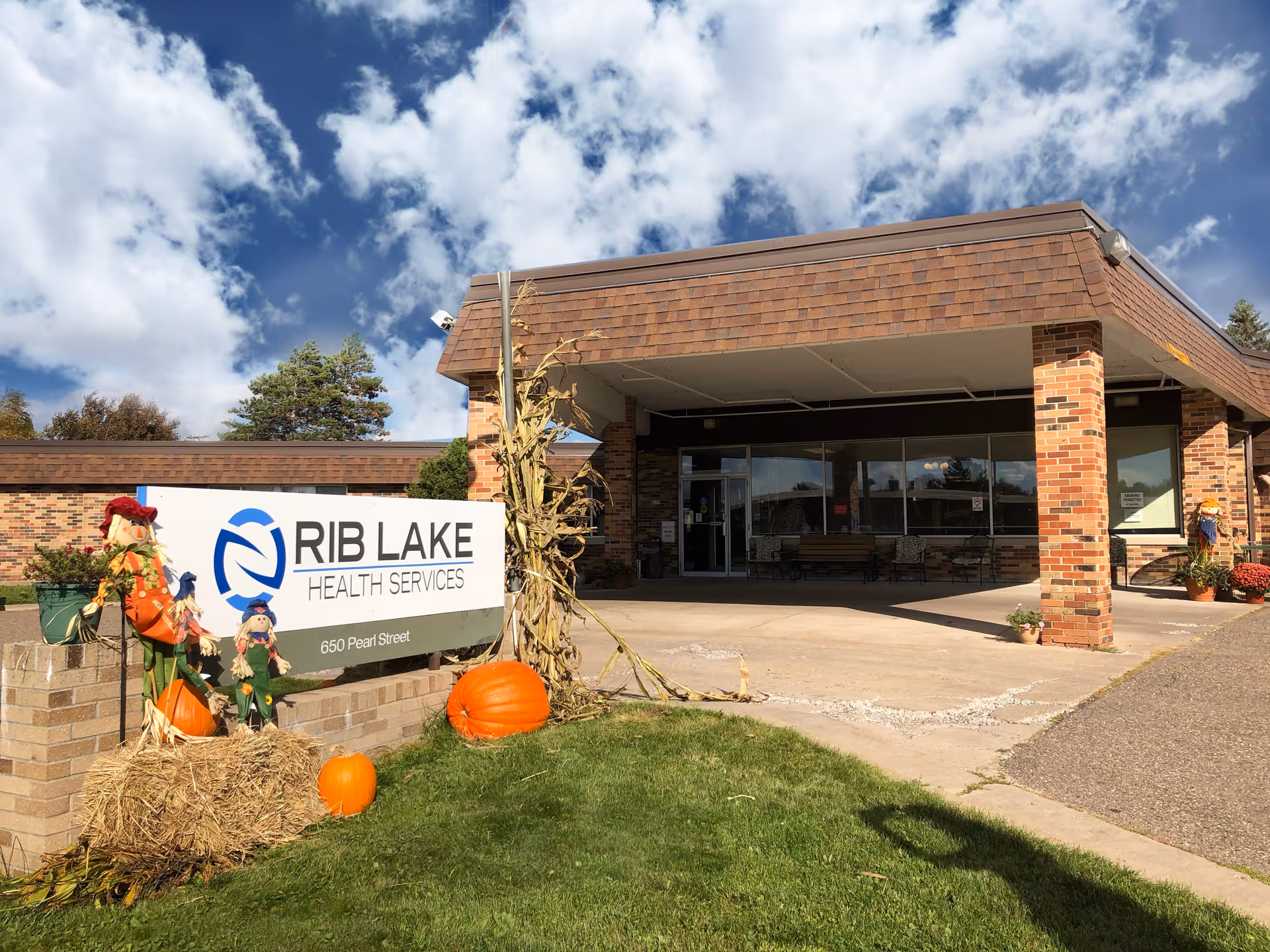 Exterior view of Rib Lake Health Services building with a covered entrance, brick pillars, and a sign displaying the facility name and address. The area is decorated with autumn-themed items including pumpkins, hay bales, and scarecrows. The sky is partly cloudy.