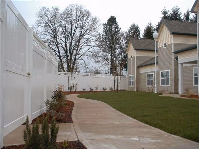 Curved concrete pathway bordered by grass and small plants leading alongside a beige building with white trim and a white fence under a cloudy sky.