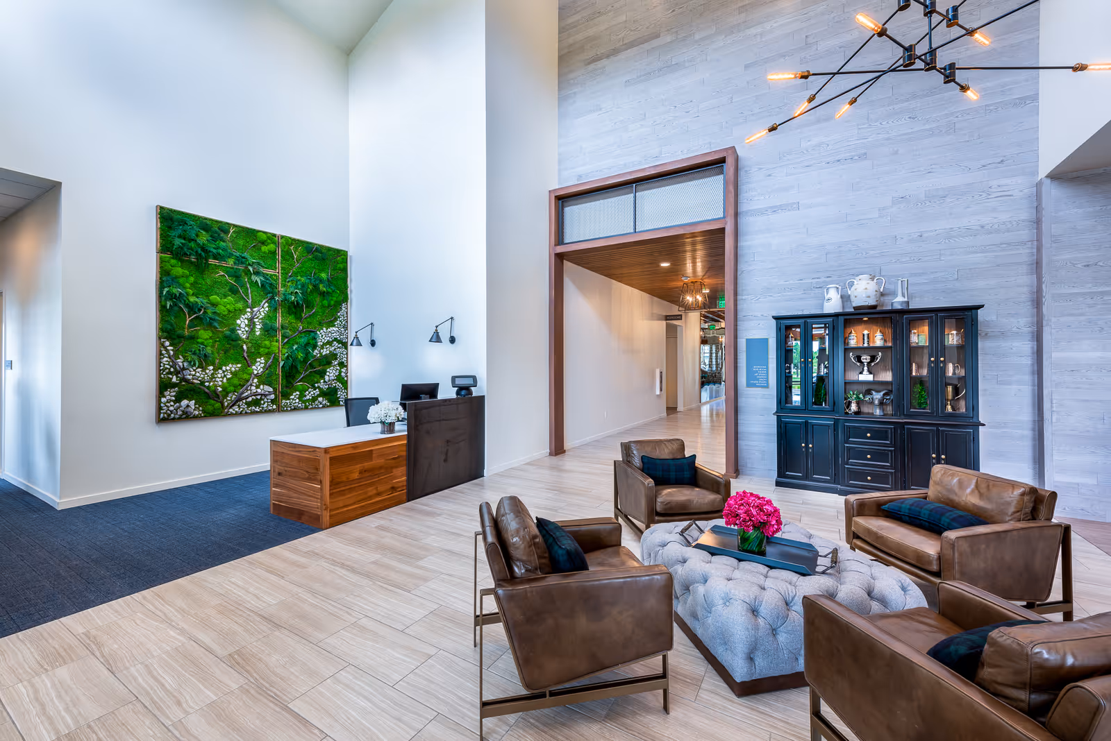 A modern and spacious lobby area with high ceilings, featuring a reception desk on the left with two wall-mounted lamps above it. In the center, there is a seating area with four brown leather armchairs arranged around a large tufted ottoman with a vase of pink flowers and books on top. On the right side, there is a black display cabinet with decorative items inside. The walls are light-colored with a large green nature-themed artwork hanging near the reception desk.