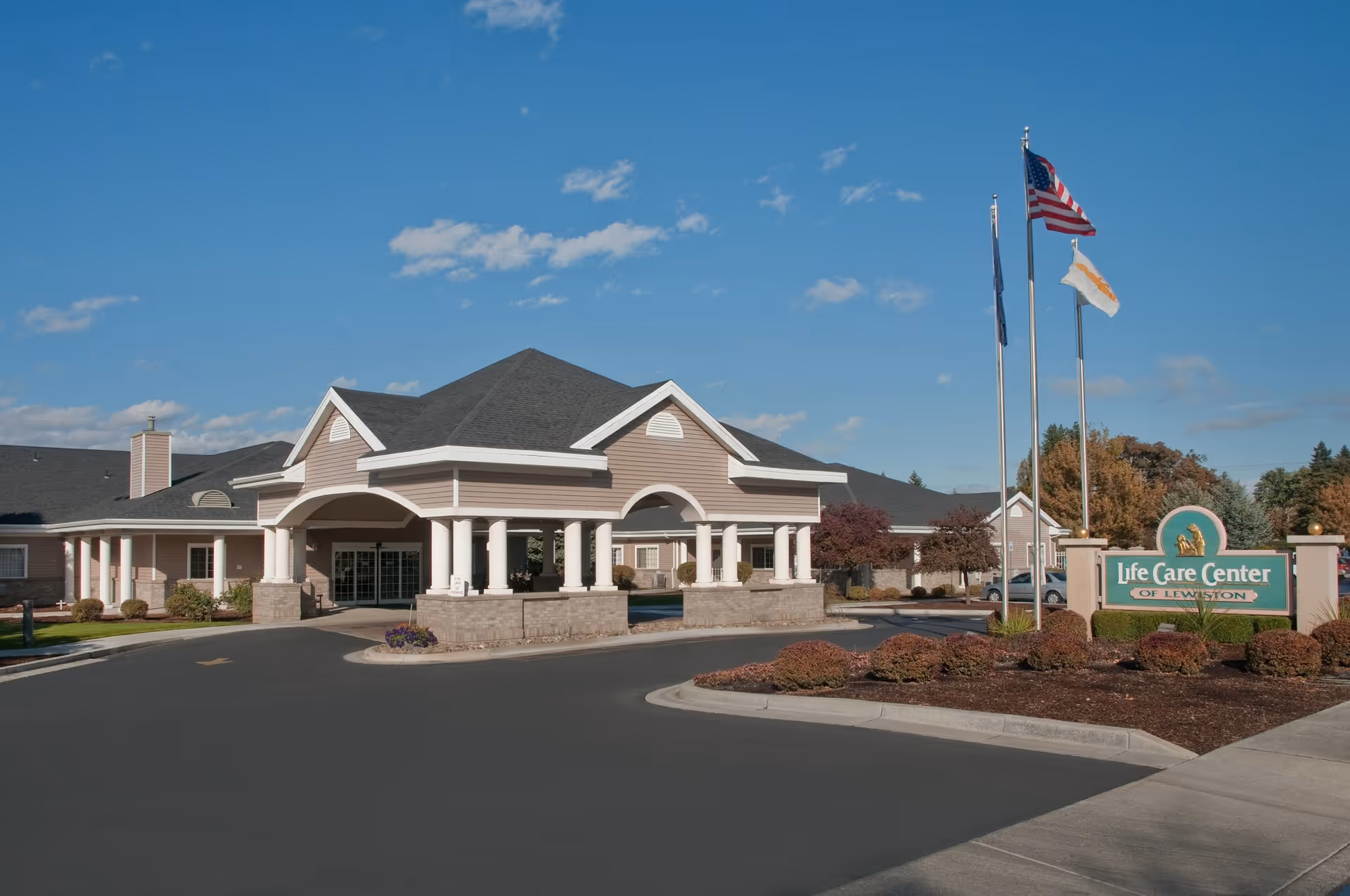 Exterior view of the Life Care Center of Lewiston building under a blue sky with some clouds. The building features a covered entrance with white columns and a dark roof. There are three flagpoles with flags, and a sign in front that reads 'Life Care Center of Lewiston' surrounded by landscaped bushes.