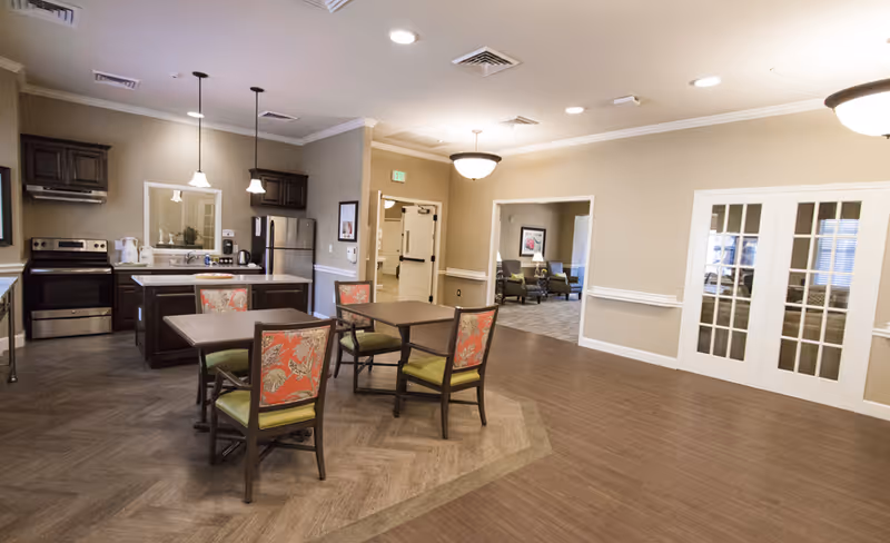Communal dining area and kitchenette featuring several tables with patterned chairs, a stainless-steel stove and refrigerator, and seating visible through doorways.