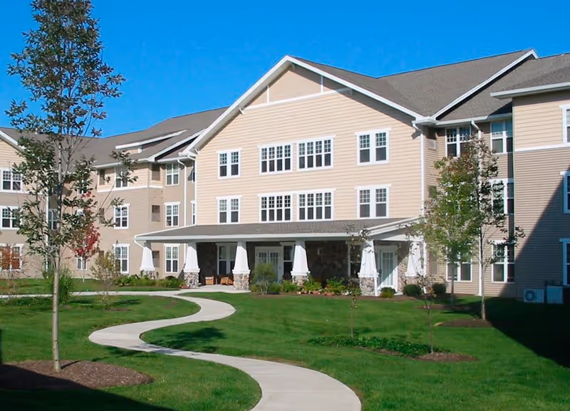 Exterior view of a multi-story senior living facility building with beige siding and white trim. The building has multiple windows and a covered entrance supported by white columns. A curved concrete walkway leads through a well-maintained green lawn with young trees and landscaped areas under a clear blue sky.