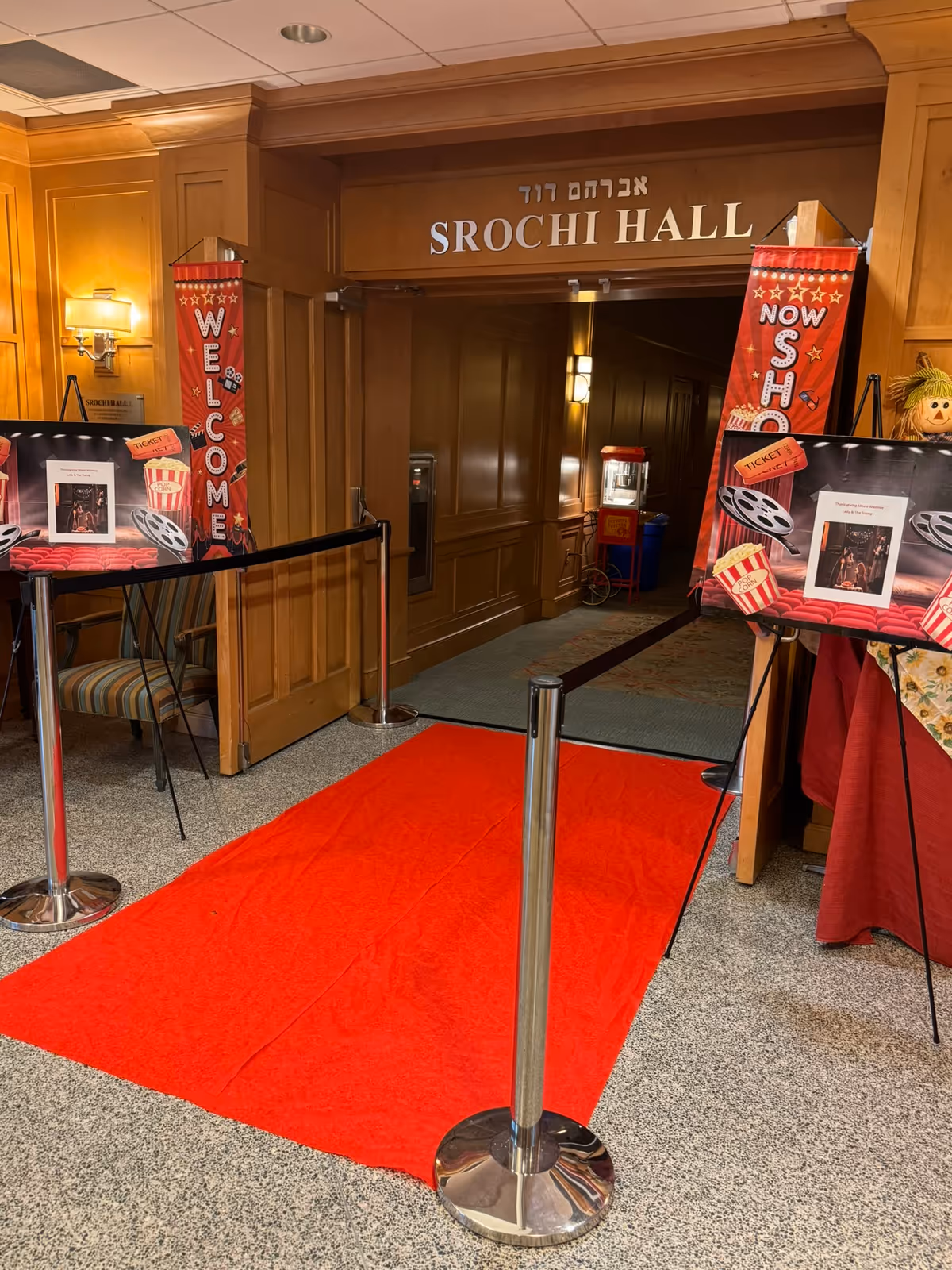 Entrance to Srochi Hall with a red carpet laid out and stanchions with black ropes on either side. There are two vertical banners on either side of the entrance, one says 'WELCOME' and the other says 'NOW SHOWING' with movie-themed decorations including popcorn and film reels. The walls and door frame are wooden, and there is a popcorn machine visible inside the hall.