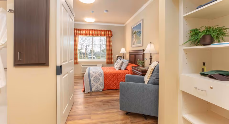A cozy bedroom in a memory care community featuring a bed with an orange quilt and patterned blanket, two bedside tables with lamps, a blue armchair, a window with red plaid curtains, a framed landscape painting on the wall, and a built-in shelving unit with a potted plant and other items.