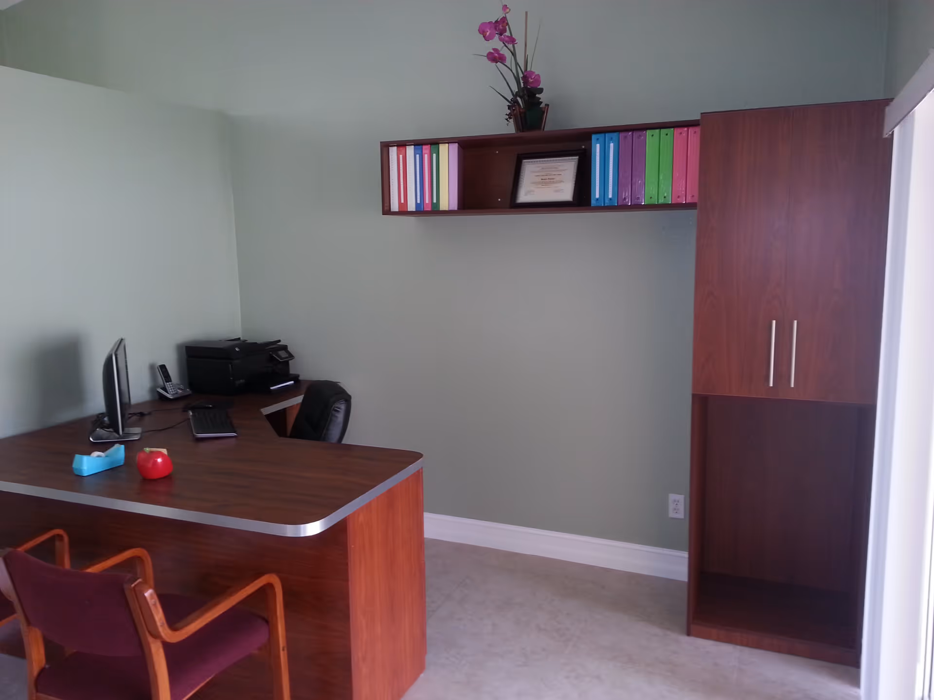 A small office space with a wooden desk and two chairs, a computer monitor, keyboard, printer, and a telephone on the desk. Above the desk is a wooden wall-mounted shelf holding colorful binders, a framed certificate, and a potted plant with pink flowers. There is also a wooden cabinet with closed doors and an open shelf on the right side of the room. The walls are painted light green and the floor is tiled.