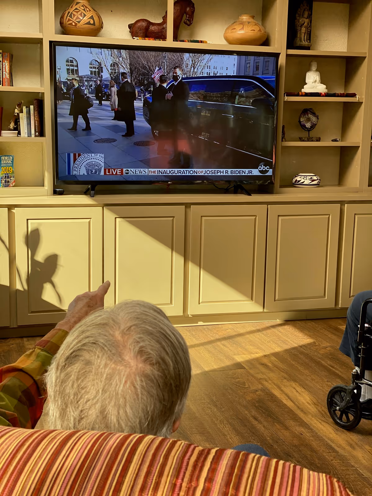 An elderly resident points toward a television in a communal living room while watching a news broadcast.