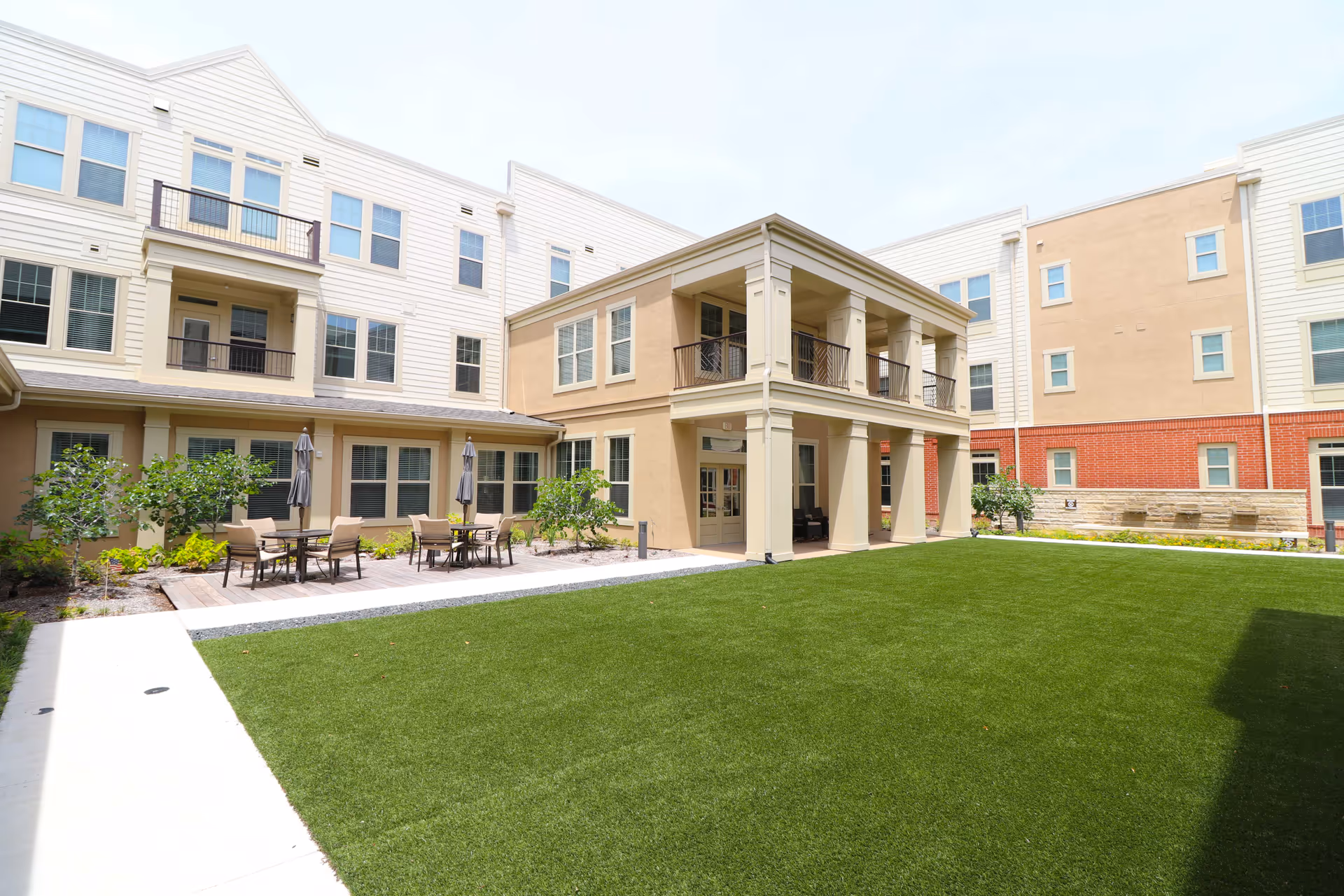 Outdoor courtyard area of Watermere at Frisco senior living facility featuring a well-maintained green lawn, patio tables with chairs and umbrellas, surrounded by a three-story building with balconies and multiple windows.