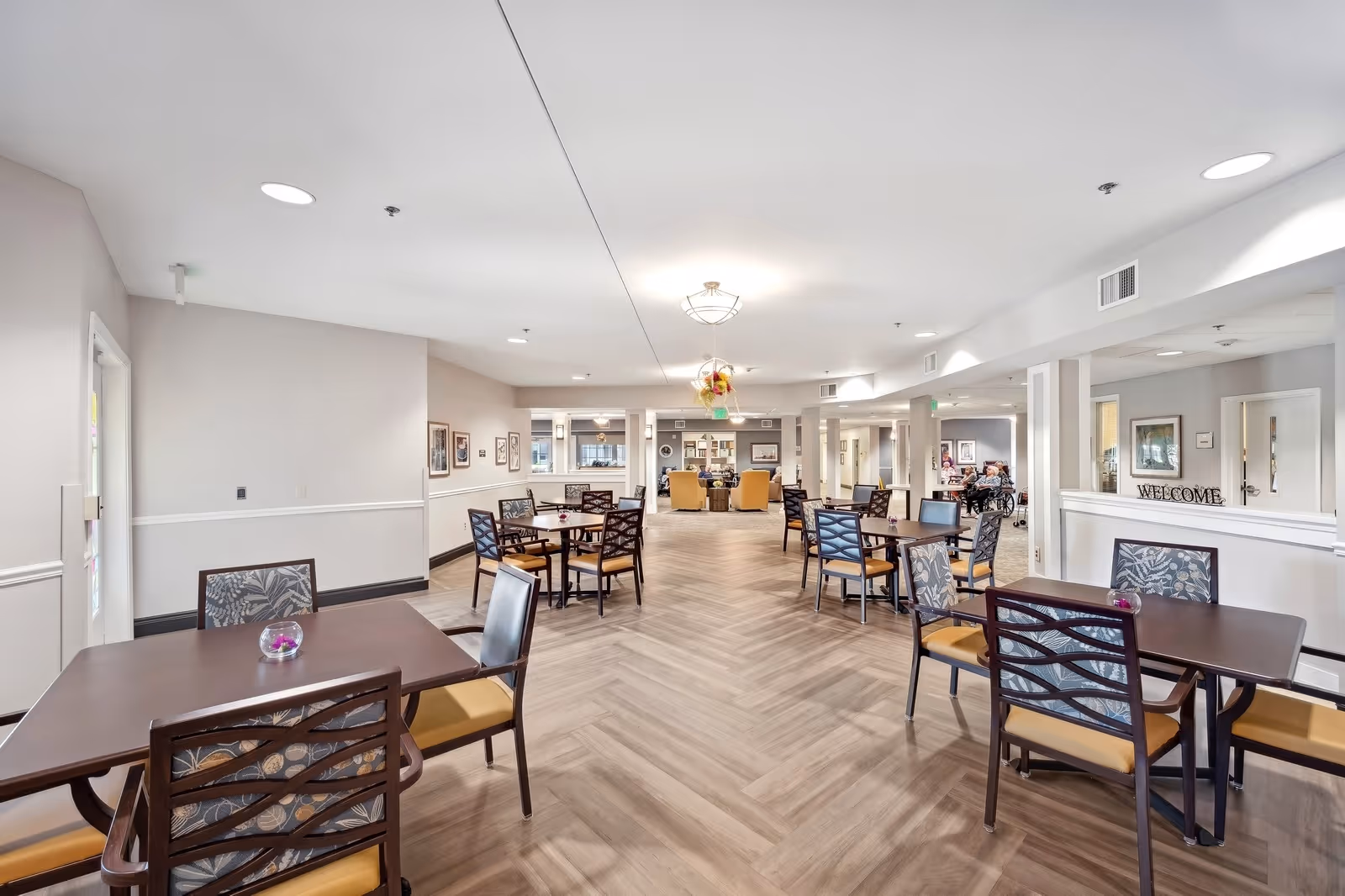 A spacious and well-lit common dining area in a senior living facility with multiple tables and chairs arranged neatly. The room features light-colored walls, wood-patterned flooring, ceiling lights, and a welcoming atmosphere. In the background, there is a seating area with armchairs and some framed pictures on the walls.
