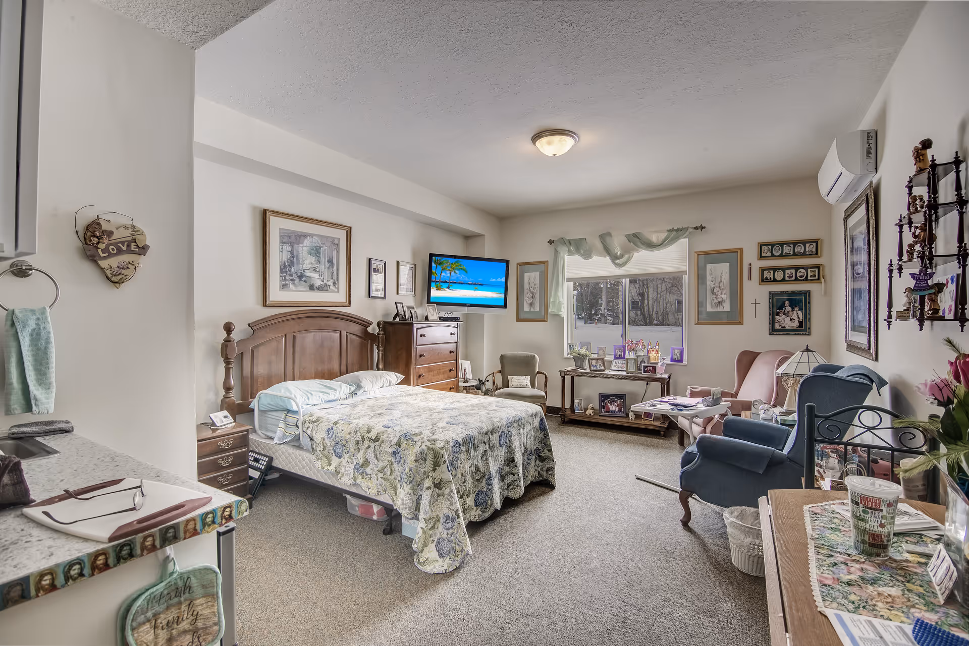 A cozy bedroom in a senior living facility with a wooden bed covered in a floral quilt, a dresser with a TV mounted above it, two armchairs, a window with sheer curtains, and various framed pictures and decorations on the walls and surfaces.