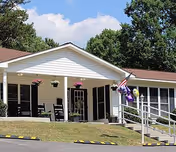 Exterior view of a single-story assisted living facility building with a covered entrance, a ramp with handrails, hanging flower pots, and an American flag. Trees and a clear sky are visible in the background.
