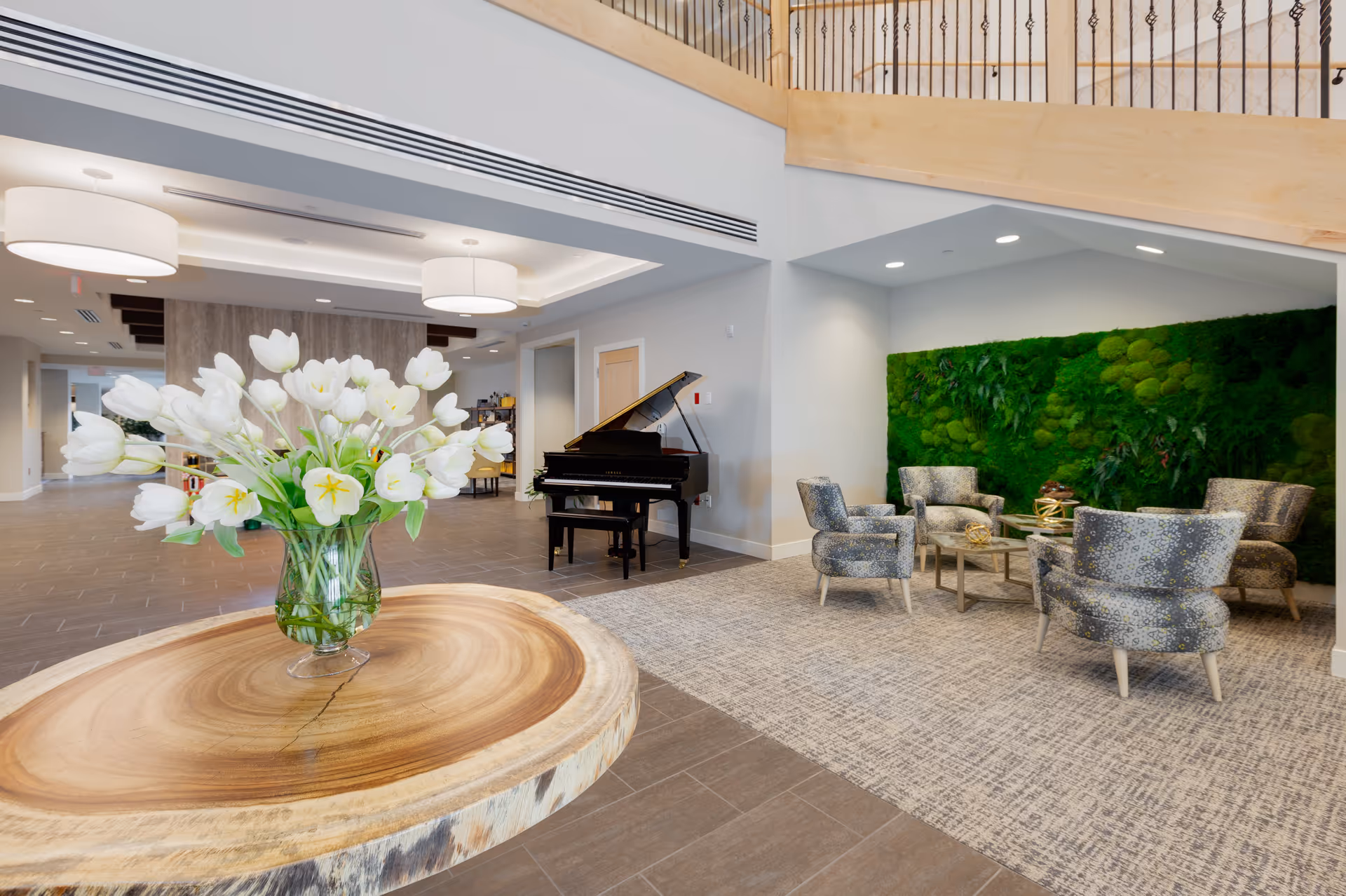 A spacious and modern living room area in Melody Living Independent Living facility featuring a large wooden round table with a glass vase of white tulips in the foreground, a black grand piano against the wall, and a seating area with four patterned armchairs around a coffee table in front of a green moss and plant wall.