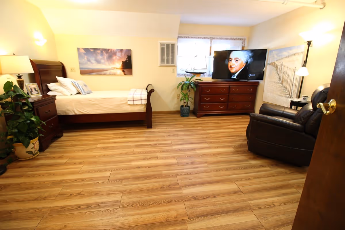 A cozy bedroom with a wooden bed, nightstand with a lamp and plant, a wooden dresser with a TV showing a portrait of a man, a leather armchair, and wall art depicting a beach scene and a sunset.
