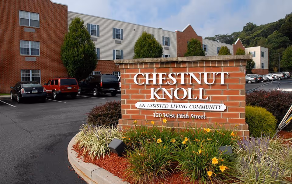 A brick sign for Chestnut Knoll, an assisted living community, located at 120 West Fifth Street. Behind the sign is a parking lot with several parked cars and a multi-story residential building with brick and beige siding. There are trees and landscaped plants around the sign and parking area.