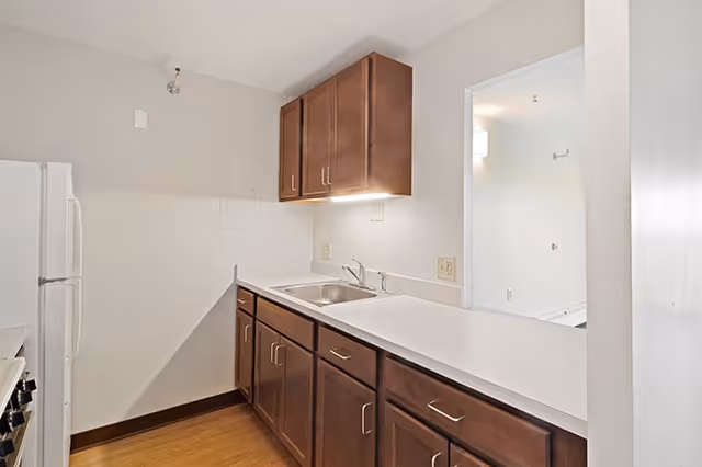 A small kitchen area with wooden cabinets, a white countertop, a stainless steel sink, and a white refrigerator. The kitchen has light-colored walls and a wooden floor. There is an open pass-through window to an adjacent room.