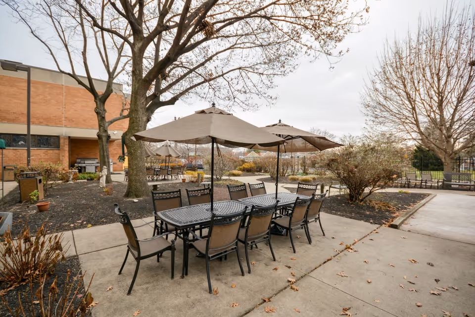 Outdoor patio area with multiple metal tables and chairs under large beige umbrellas. The area is surrounded by leafless trees and shrubs, with a brick building visible on the left side. The ground is paved with concrete, and there are some fallen leaves scattered around.