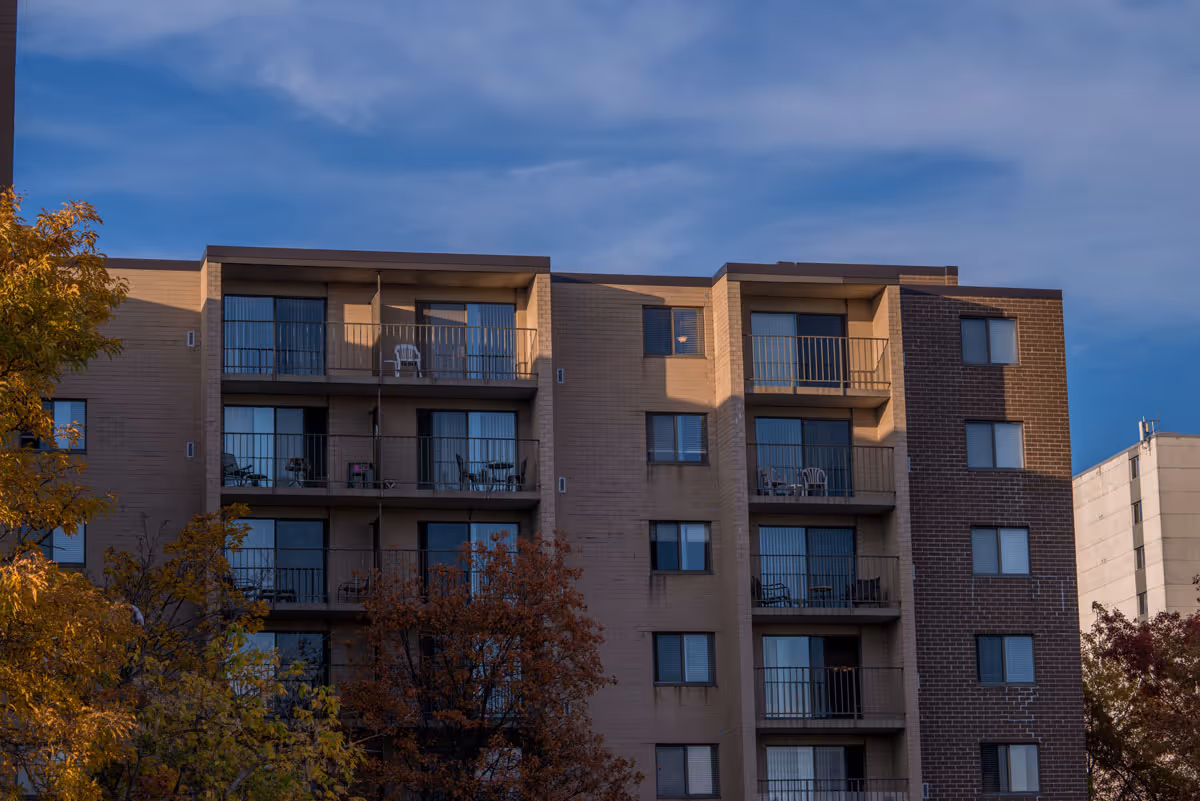 Exterior view of a multi-story senior living facility building with balconies on each floor. The building has a mix of light and dark brick walls. Trees with autumn foliage are visible in the foreground under a partly cloudy blue sky.
