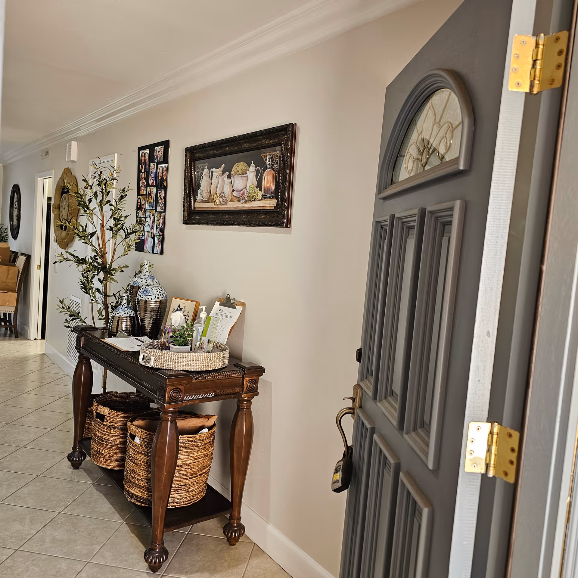 View of an open gray front door leading into a hallway with a wooden console table against a white wall. The table holds decorative items including two large blue and white vases, a small plant, hand sanitizer, and a clipboard. Below the table are two woven baskets. The wall above the table features framed artwork and a photo collage. The floor is tiled in a light color.
