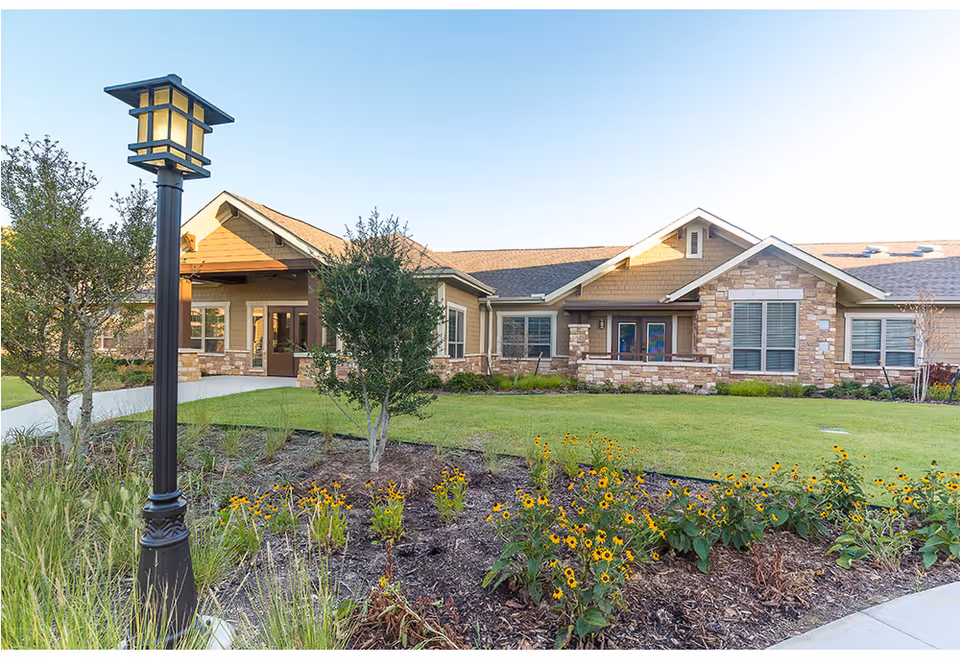 Exterior view of a single-story senior living facility building with stone and wood siding, surrounded by a well-maintained lawn, flower beds with yellow flowers, small trees, and a black lamp post in the foreground under a clear blue sky.