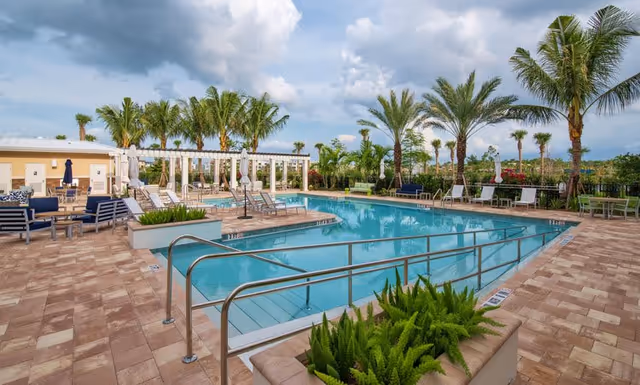 Outdoor swimming pool with lounge chairs, palm trees, and a paved deck under a partly cloudy sky.