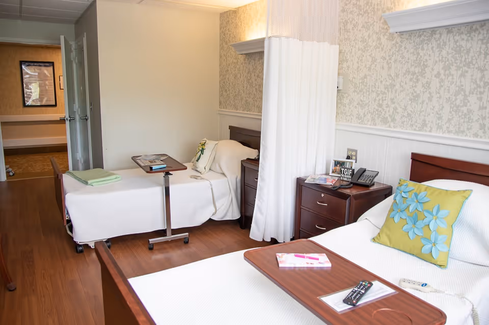 A nursing facility room with two hospital-style beds separated by a white privacy curtain. Each bed has a wooden headboard, white bedding, and a small overbed table with items like a remote control and a book. There are two wooden nightstands between the beds with a telephone, framed photos, and magazines. The room has wood flooring and light-colored wallpaper with a subtle floral pattern.