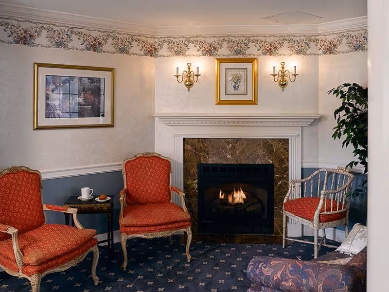 A cozy sitting area with a lit fireplace surrounded by a marble frame. There are two red upholstered armchairs with wooden frames, a small side table with a cup and plate, a white wooden chair with a red cushion, and a patterned sofa partially visible. The walls have floral wallpaper border near the ceiling, two wall sconces with lights, and framed artwork. A potted plant is in the corner.
