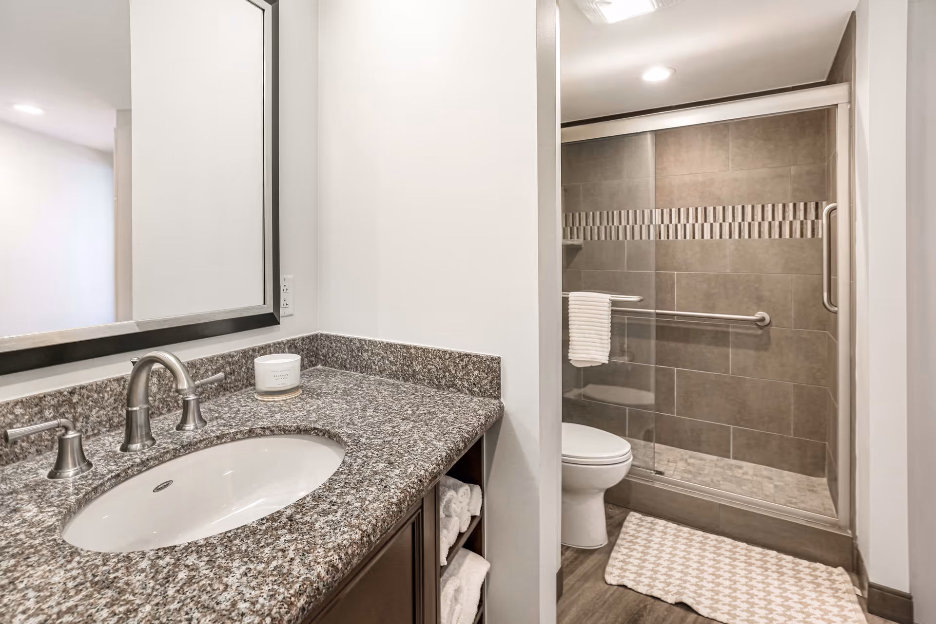 A modern bathroom featuring a granite countertop with an oval sink and silver faucet. A large mirror is mounted above the sink. To the right, there is a walk-in shower with glass sliding doors, gray tiles, and a decorative tile strip. A white towel hangs on a grab bar inside the shower, and a white toilet is positioned next to the shower. The floor has a patterned bath mat in front of the shower.