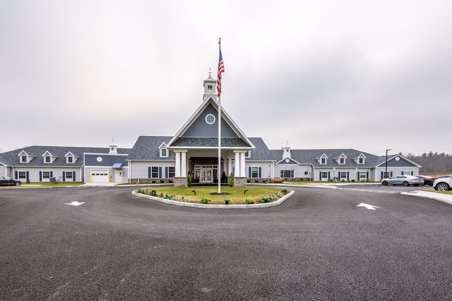 Front exterior view of Peregrine Senior Living at Colonie, featuring a large building with gray siding, white trim, multiple windows, and a central entrance with a covered porch. An American flag is prominently displayed on a flagpole in front of the building, surrounded by a landscaped circular driveway with parked cars on either side.