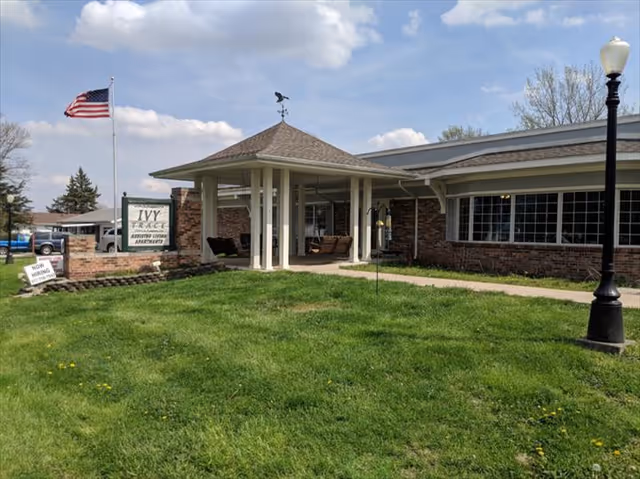 Exterior view of Ivy Trace Assisted Living Apartments building with a covered entrance, an American flag on a flagpole, a green lawn, and a street lamp in front.