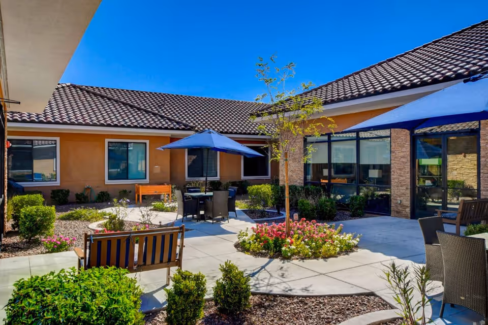Outdoor courtyard area at Grand Montecito Memory Care with patio seating including chairs and tables shaded by blue umbrellas, surrounded by landscaped plants, flowers, and small trees, with a building featuring tiled roof and large windows in the background under a clear blue sky.