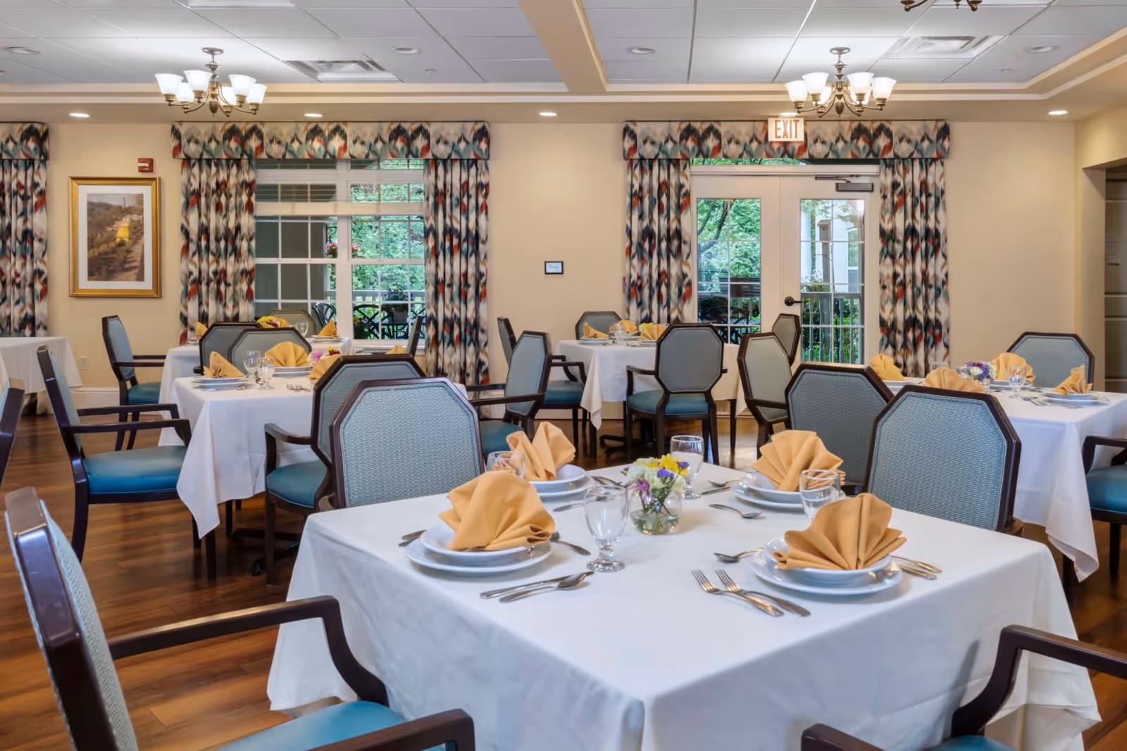 Bright dining room with multiple tables set with white linens, folded napkins, place settings and chairs.