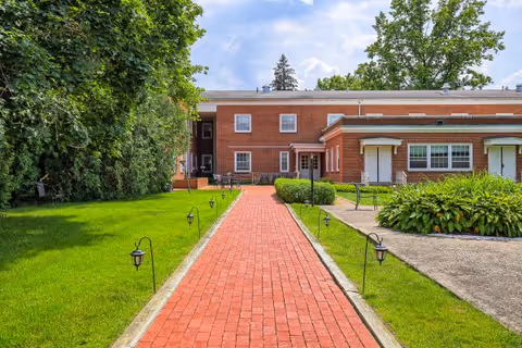 A red brick walkway lined with small garden lights leads to the entrance of a two-story brick building. The building is surrounded by green grass, bushes, and trees under a partly cloudy sky.