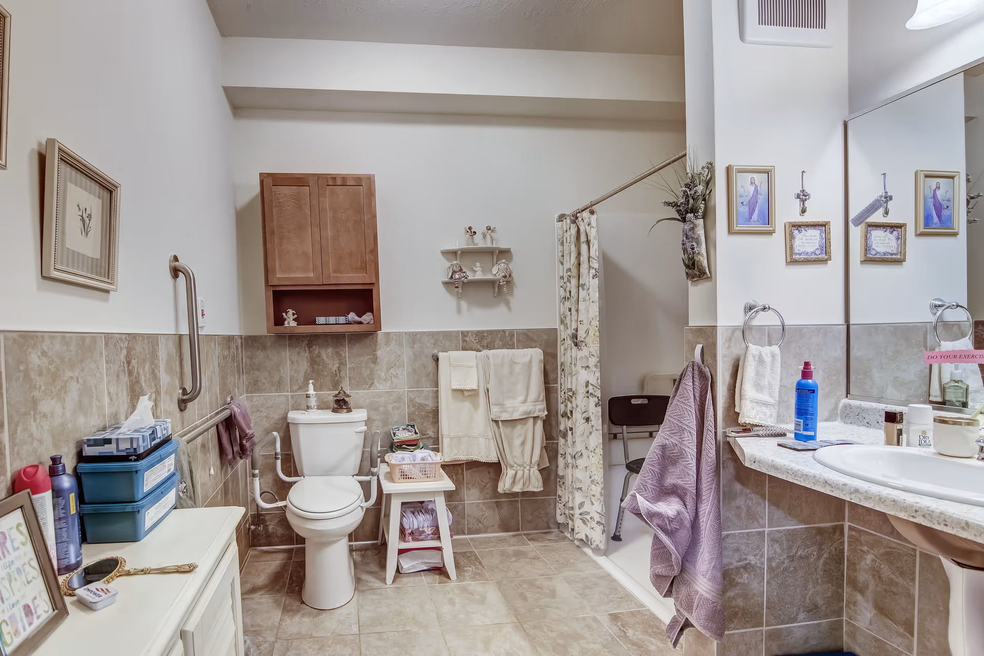 A bathroom with beige tiled walls and floor, featuring a toilet with support rails, a small white stool with folded towels, a shower area with a floral curtain and a black shower chair, a sink with a large mirror above it, and various toiletries and decorations including framed pictures and hanging towels.