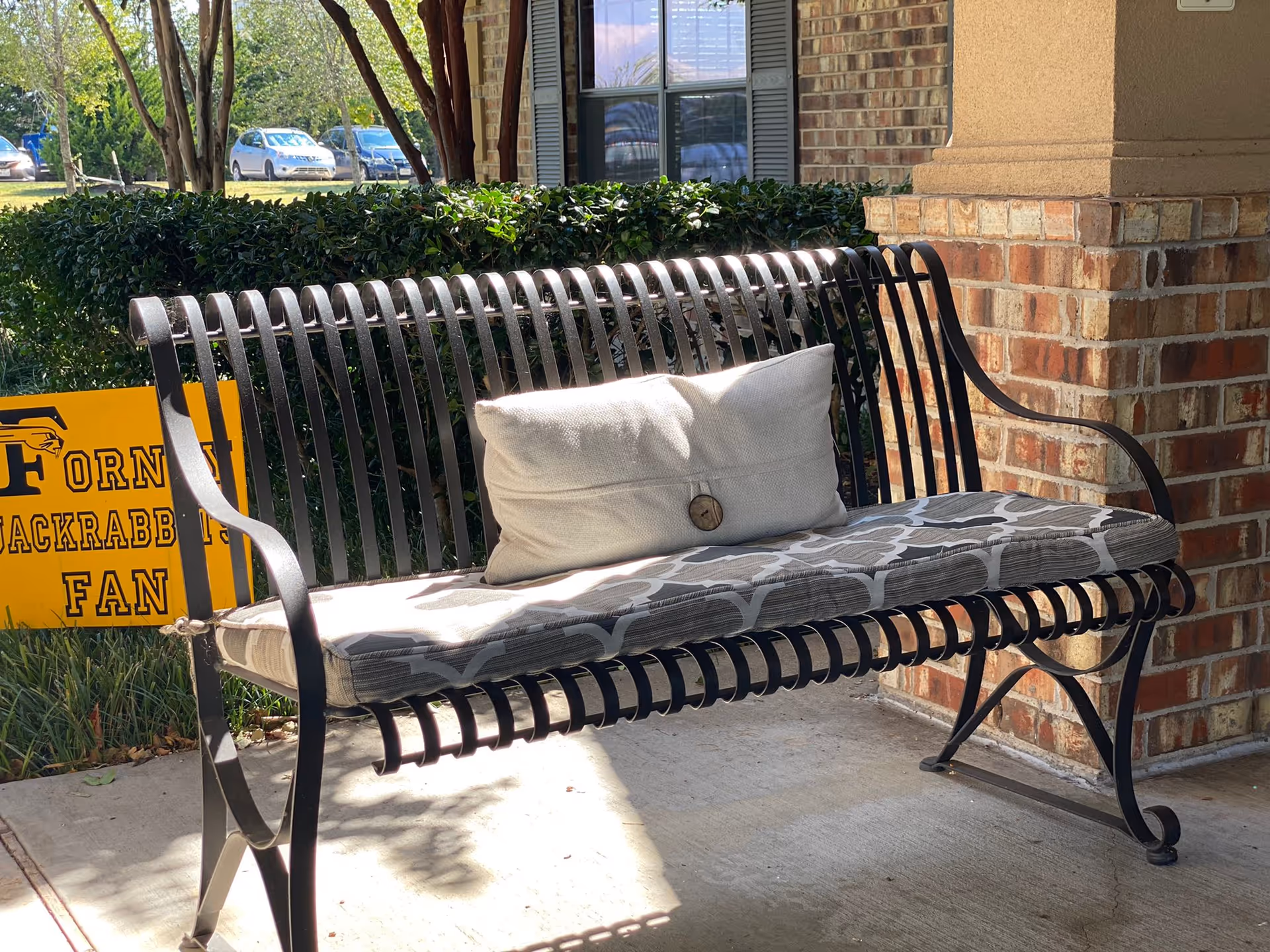 A black metal outdoor bench with a patterned cushion and a white pillow with a button is placed on a concrete porch next to a brick wall. Behind the bench, there is a green hedge and a yellow sign that reads 'FORNEY JACKRABBITS FAN'. Several parked cars and trees are visible in the background.