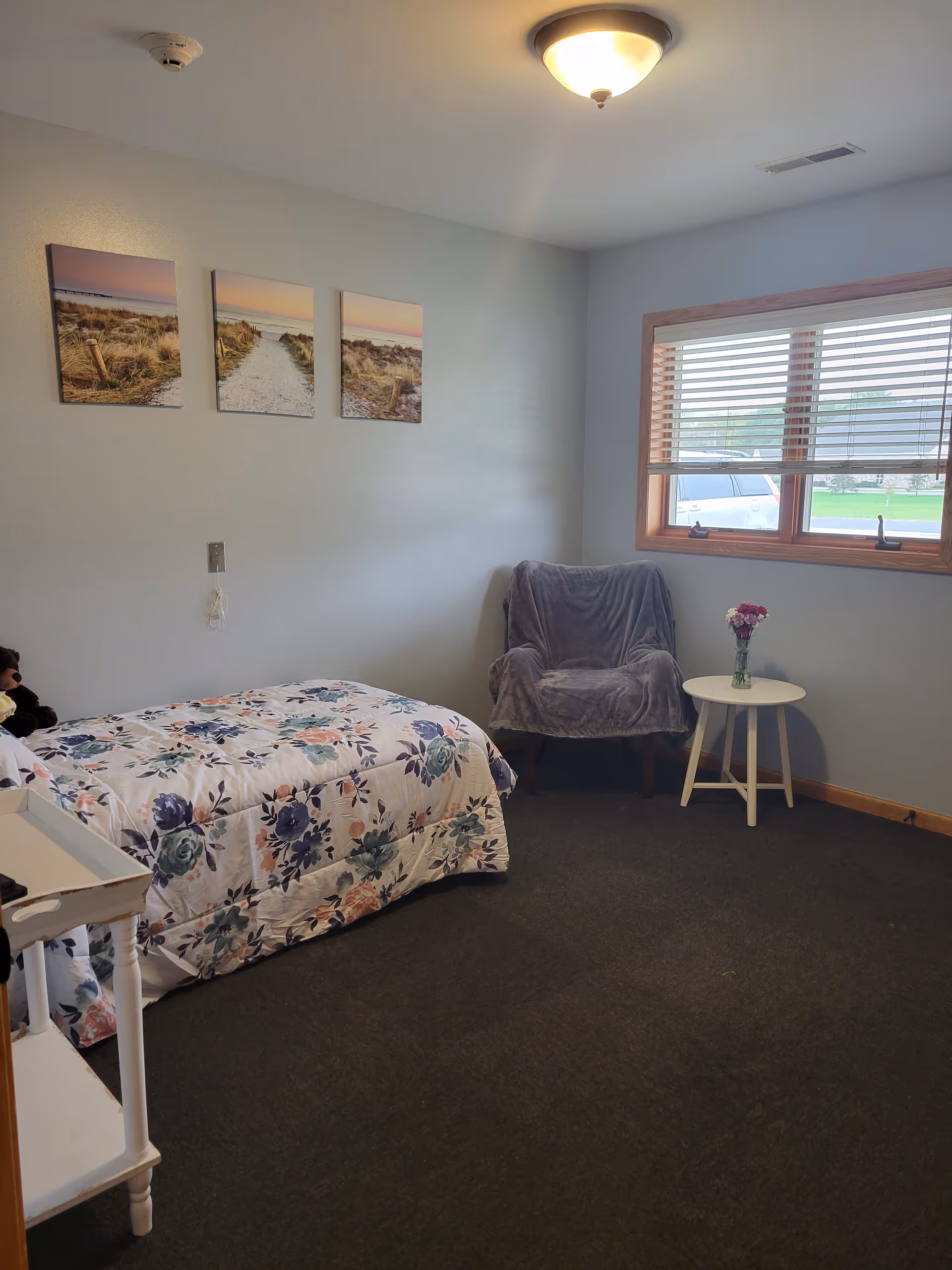 A small bedroom with a single bed covered in a floral comforter, a gray armchair draped with a gray blanket, a small white round table with a vase of flowers, a window with blinds, and three landscape pictures on the wall.