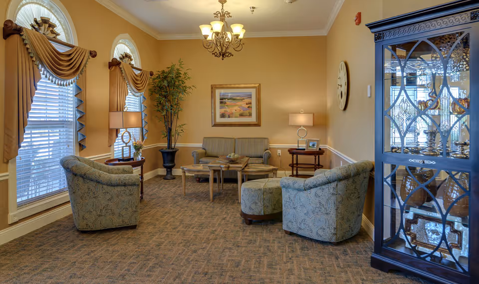 A cozy senior living facility lounge area with two patterned armchairs, a matching ottoman, a striped loveseat, and two wooden side tables with lamps. The room has beige walls, carpeted floor, two large arched windows with decorative curtains, a chandelier, a large potted plant, a framed landscape painting, a wall clock, and a glass-front cabinet displaying decorative items.