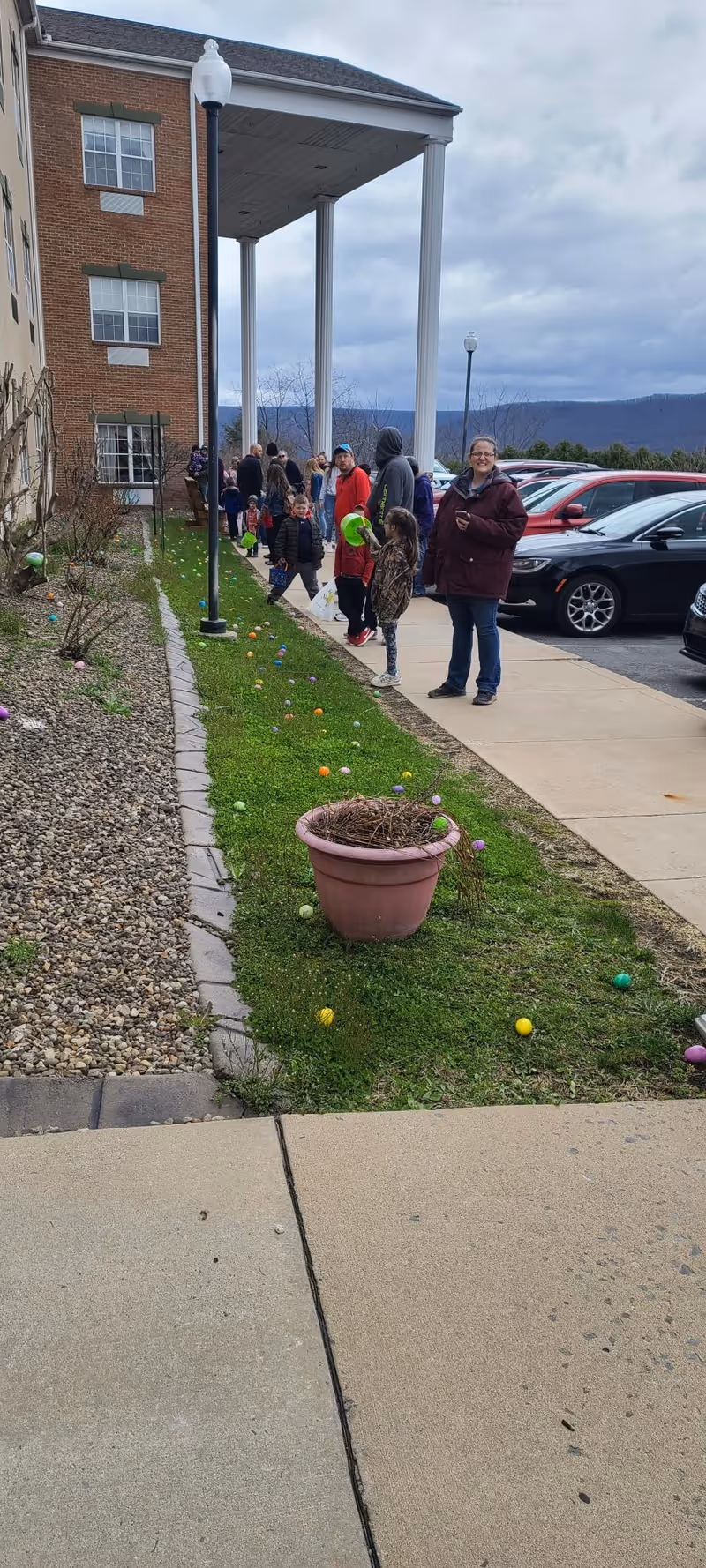 People, including children and adults, standing and walking along a sidewalk next to a brick building with large white columns. The grassy area beside the sidewalk is scattered with colorful plastic Easter eggs. Several parked cars are visible on the right side under a cloudy sky.