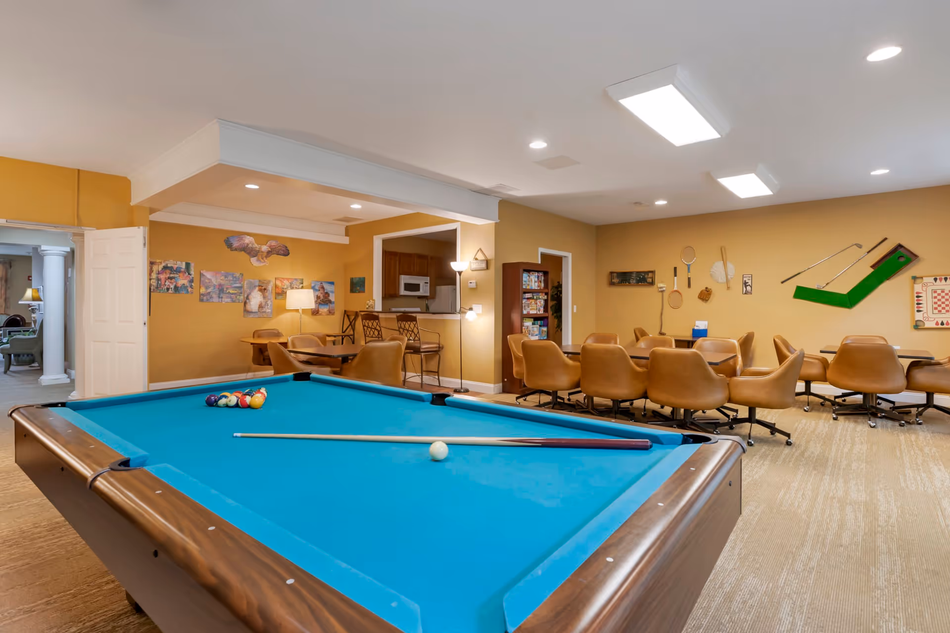 A recreational room with a pool table in the foreground and a cue stick resting on it. Behind the pool table, there are several tables with brown swivel chairs arranged around them. The walls are decorated with various game-related items including tennis rackets, golf clubs, and a checkerboard. There is a small kitchen area with a microwave and bar stools, and a bookshelf filled with board games. The room has warm yellow walls and bright ceiling lights.