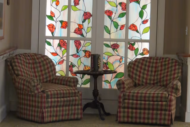 Two plaid upholstered armchairs with red, green, and beige colors are positioned on either side of a small round wooden table. Behind them are double doors with stained glass panels featuring a floral design with red flowers and green leaves.