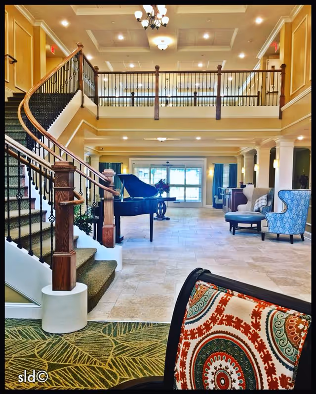 Interior view of a senior living facility lobby featuring a grand staircase with wooden handrails and black iron balusters on the left. In the center, there is a black grand piano and a round table with a flower arrangement. The floor is tiled, and there are several upholstered chairs and a small sofa arranged on the right side. The ceiling has recessed lighting and chandeliers, and large windows with blue curtains are visible in the background.