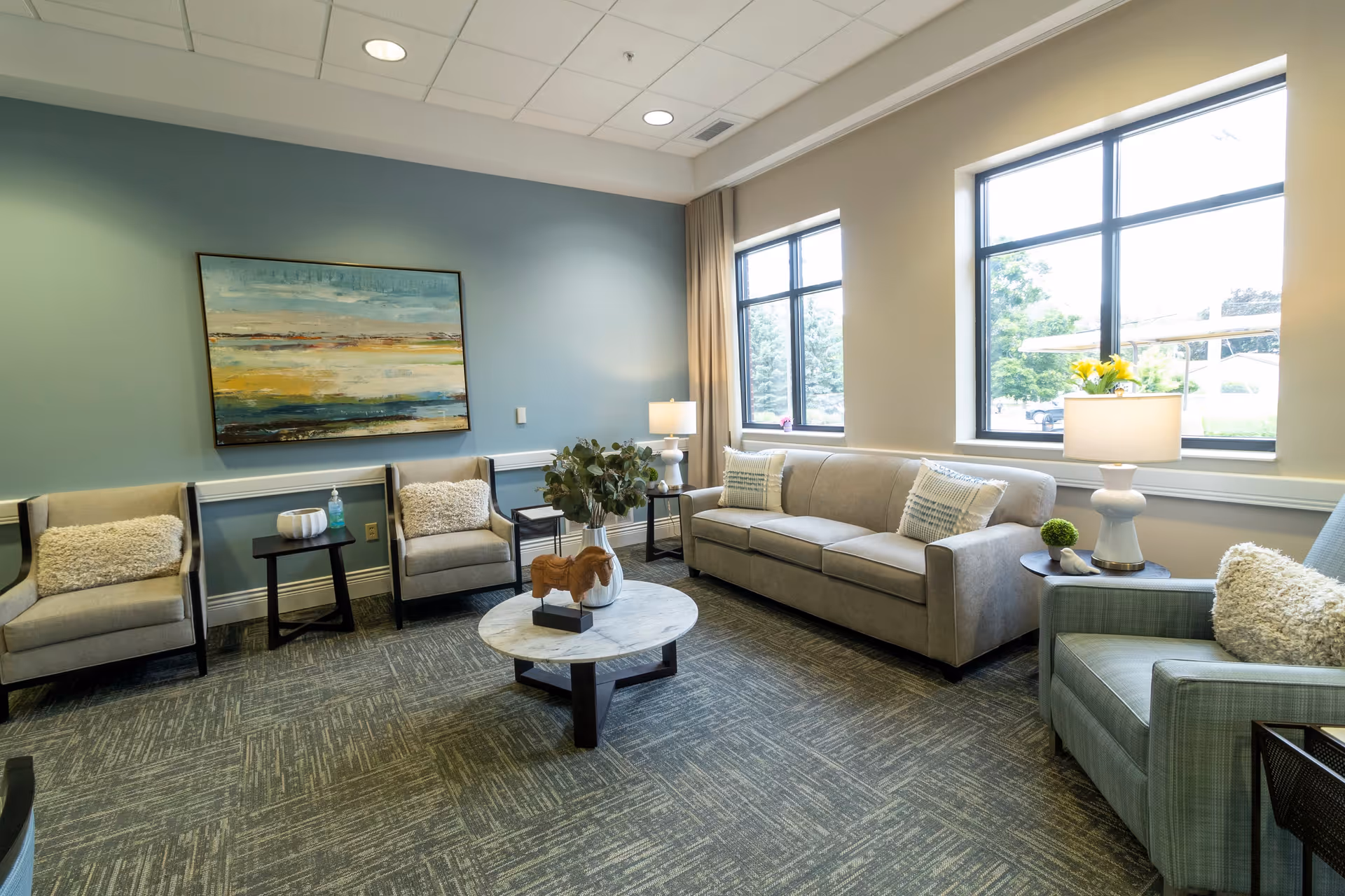 A bright and cozy living room with a beige sofa, two beige armchairs, and a green armchair, all with decorative pillows. A round marble coffee table with a wooden horse sculpture and a vase with greenery sits in the center. Two large windows let in natural light, and a modern painting hangs on the blue accent wall. There are two side tables with lamps and small decorative items.