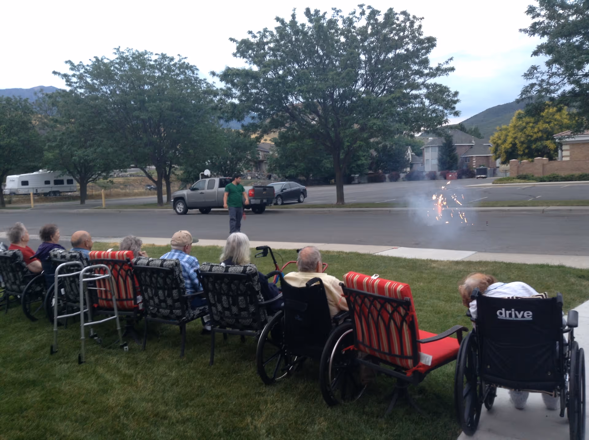 A group of elderly people sitting outdoors on chairs and wheelchairs on a grassy area, watching small fireworks on the street. A man stands nearby observing the fireworks. Trees, parked cars, and houses are visible in the background under a cloudy sky.