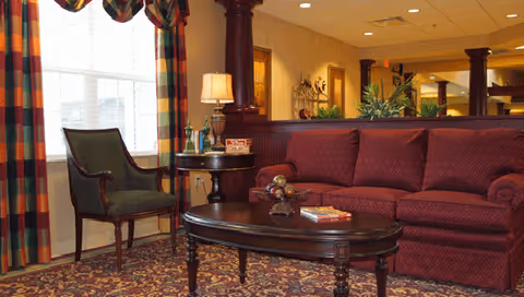 A cozy living room area with a maroon sofa, a dark wooden coffee table with decorative items and a book, a side table with a lamp and bottles, a green upholstered chair, patterned curtains, and a patterned carpet. The room has warm lighting and decorative plants in the background.