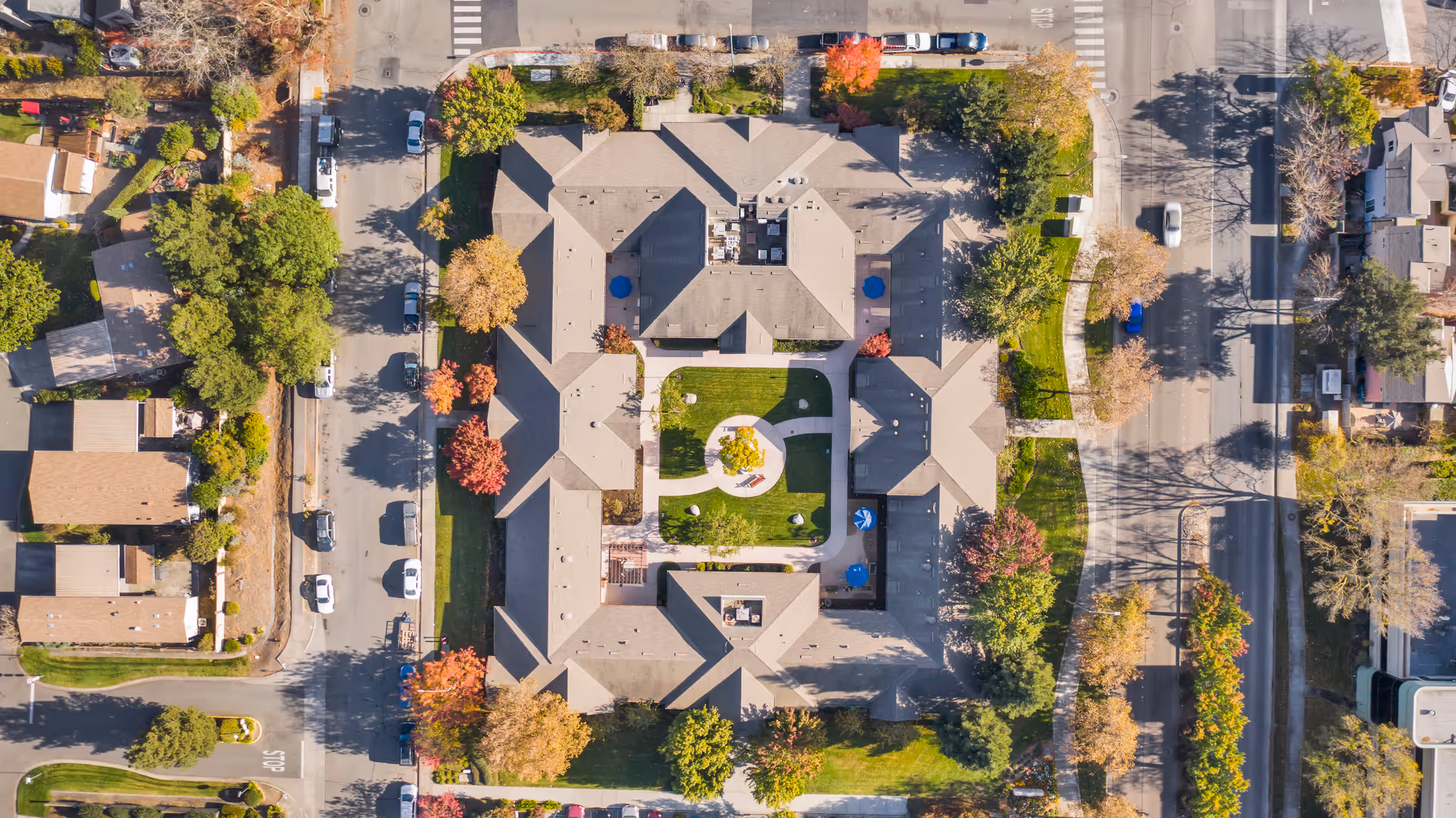 Aerial view of MuirWoods Memory Care facility showing a large building with a central courtyard featuring green grass, trees, and walking paths. Surrounding the building are streets with parked cars and additional trees with autumn foliage.