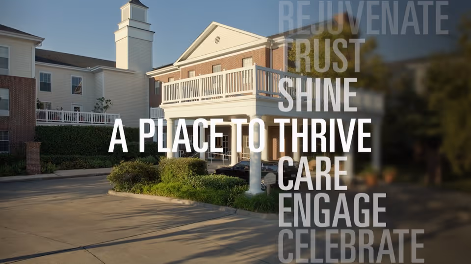 Exterior view of Belmont Village Senior Living West University building with a covered entrance, white columns, brick and white siding, surrounded by greenery and a clear sky.