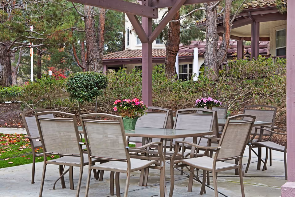 Outdoor patio area with a glass-top table surrounded by eight beige mesh chairs under a wooden pergola. There are two flower pots with pink flowers on the table. In the background, there are trees, bushes, and a building with a tiled roof and windows.